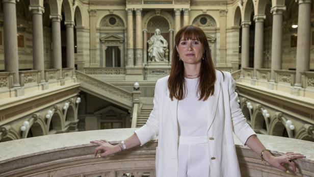Frau in weißem Anzug steht in einer prunkvollen Halle mit Säulen und Treppe.