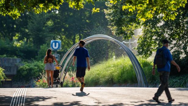 Menschen gehen auf einem sonnigen Weg im Park, ein Mann schiebt einen Kinderwagen, daneben ist ein U-Bahn-Schild.