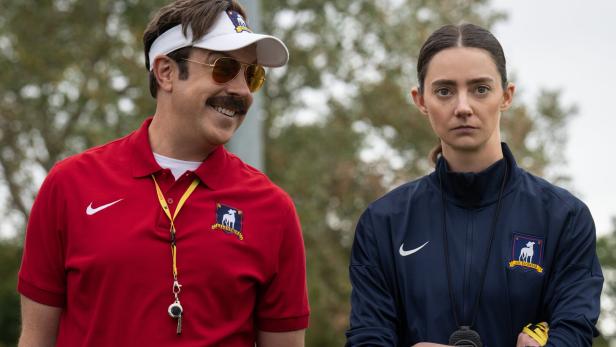 Ted Lasso mit Sonnenbrille und Visor lächelt neben der ernst dreinblickenden Trainerin Beard auf dem Trainingsgelände des AFC Richmond.