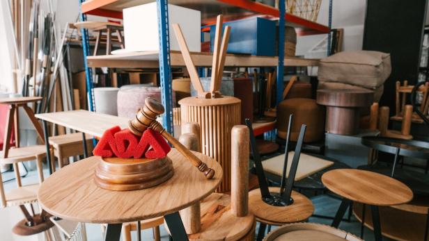 Ein Lager voller gestapelter Holzmöbel und Stühle, vorne ein Holzhammer auf einem Tisch mit roten ADA-Buchstaben.