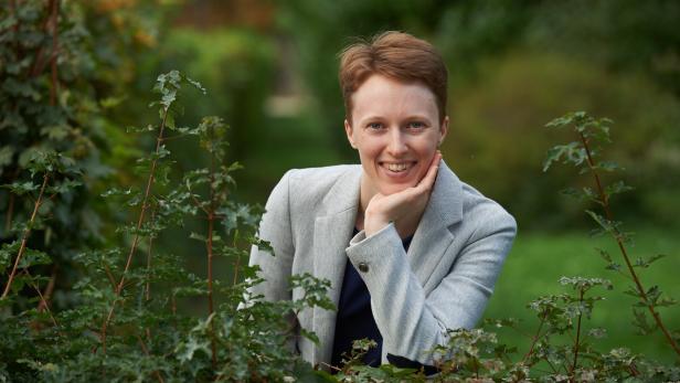 Eine kurzhaarige Frau mit hellem Blazer stützt lächelnd ihr Kinn auf die Hand, während sie hinter einem grünen Busch im Freien posiert.