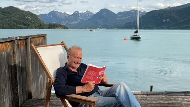 Ein Mann liest entspannt auf einer Holzterrasse am See, mit Segelboot und Bergen im Hintergrund.
