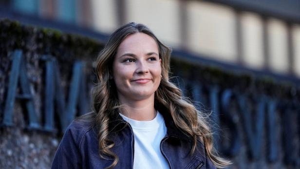 Ingrid Alexandra mit langem, gewelltem Haar trägt eine dunkle Jacke über einem weißen Shirt und lächelt vor einer steinernen Wand.