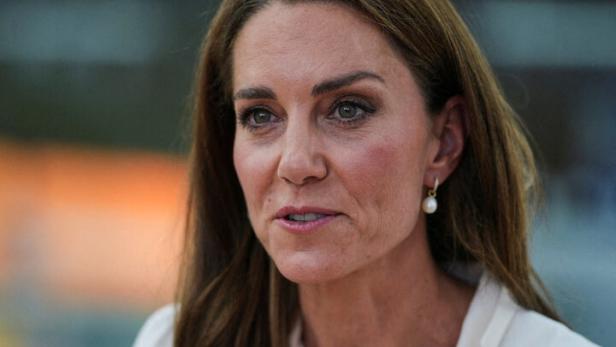 Prinzessin Kate spricht gestikulierend in einem weißen Blazer mit Perlenohrringen.