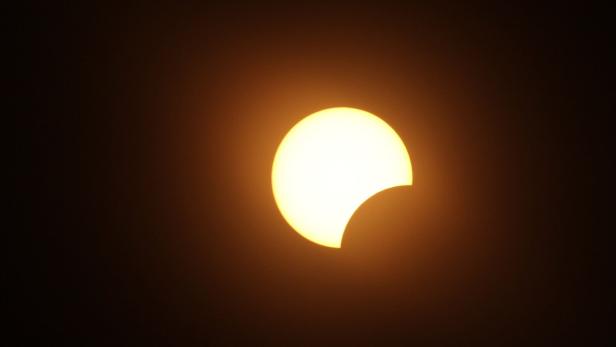 Teilweise verfinsterte Sonne mit dunkler Sichel vor orangefarbenem Himmel.