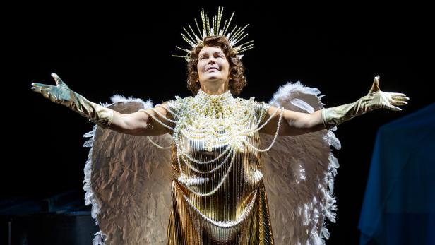 Frau in goldener Engelsrobe mit Flügeln und erhobenen Armen auf dunkler Bühne.