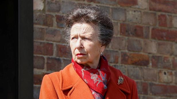 Prinzessin Anne trägt einen orangefarbenen Mantel und ein gemustertes Tuch vor einer Backsteinmauer.