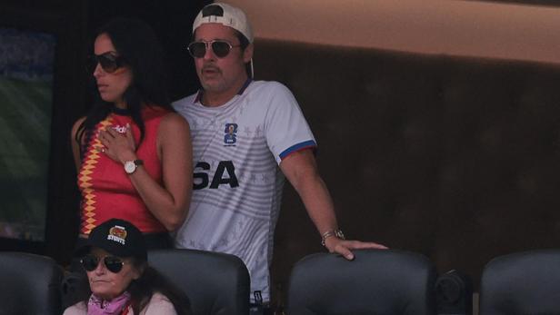 Brad Pitt trägt eine Sonnenbrille und ein USA-Trikot, während er neben einer Frau in einem roten Oberteil auf einer Tribüne steht.