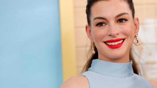 Anne Hathaway lächelt mit rotem Lippenstift und trägt ein hellblaues Oberteil vor einem blau-beigen Hintergrund.
