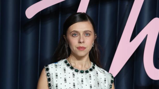 Bel Powley trägt ein weißes, ärmelloses Kleid mit blauem Blumenmuster und grünen Schmucksteinen vor einem dunklen Hintergrund.