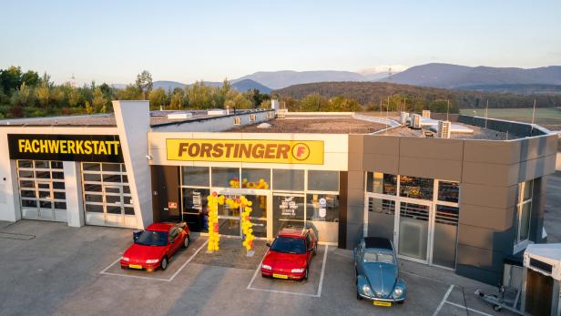 Vor einem Autohaus mit den Schildern „FACHWERKSTATT“ und „FORSTINGER“ stehen drei Autos, davor ein gelber Ballonbogen.