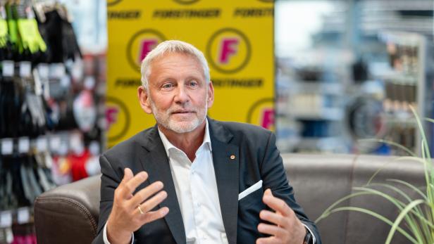 Älterer Mann im Anzug spricht gestikulierend auf einem Sofa in einem Laden, mit gelbem F‑Hintergrund.
