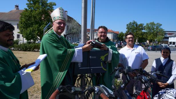 Ein Bischof segnet ein Fahrrad-Ladegerät, umgeben von Helfern und Fahrrädern im Freien.
