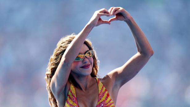 Shakira trägt eine glitzernde, gelb-rote Kleidung und formt mit ihren Händen ein Herz über ihrem Kopf.