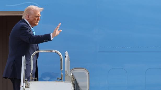 Donald Trump winkt auf einer Flugzeugtreppe neben einem blauen Regierungsflugzeug.