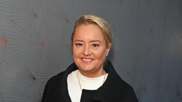 Lucy Davis lächelt in schwarzem Mantel vor einer grauen Wand.