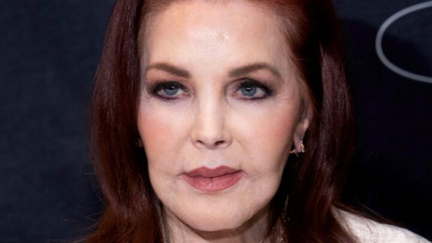 Priscilla Presley mit rötlichem Haar und weißem Blazer vor dunklem Hintergrund.