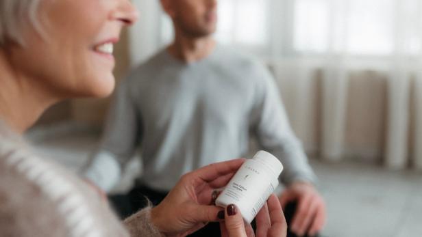 Frau hält BIOGENA Osteo Calbon Komplex Gold Dose in der Hand, Mann sitzt im Hintergrund im Schneidersitz.