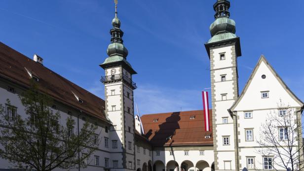 Historischer Innenhof mit zwei Türmen, Arkaden und Menschen unter blauem Himmel.