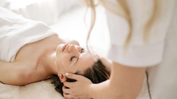 Eine Kundin liegt entspannt bei einer Kopfhautmassage im Spa, während eine Behandlerin ihren Kopf hält.