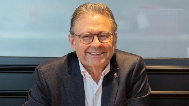 Alexander Wrabetz lächelnd im Anzug mit Brille sitzt an einem Tisch und schaut in die Kamera.