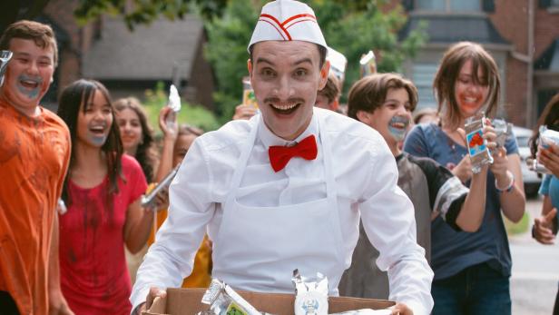 Ein Mann im weißen Dienstoutfit mit roter Fliege verteilt Süßigkeiten, umringt von jubelnden Kindern.