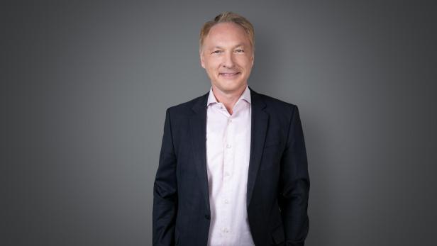 Gerhard Valeskini im dunklen Anzug und hellen Hemd steht vor grauem Hintergrund.