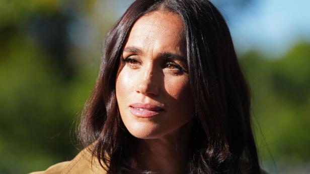 Meghan Markle blickt mit ernstem Ausdruck vor einem unscharfen, grünen Hintergrund in die Ferne.