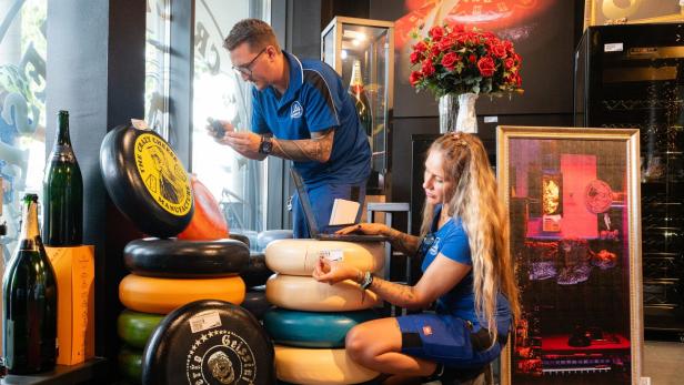 Zwei Personen in blauer Arbeitskleidung inventarisieren in einem Geschäft mehrere farbige Käselaibe von The Crazy Cheese Manufacture.