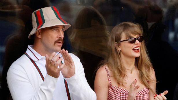 Travis Kelce mit Fischerhut und Taylor Swift in einem rot-weiß karierten Kleid klatschen gemeinsam auf einer Tribüne.