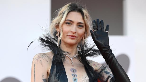 Paris Jackson winkt in einem schwarzen Lederkleid mit Federkragen und sichtbaren Tattoos auf dem roten Teppich.