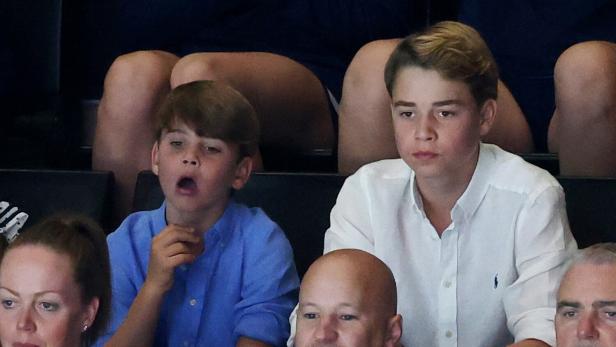 Prinz George und Prinz Louis sitzen bei einer Veranstaltung auf der Tribüne, wobei Louis gähnt und George aufmerksam nach vorne blickt.