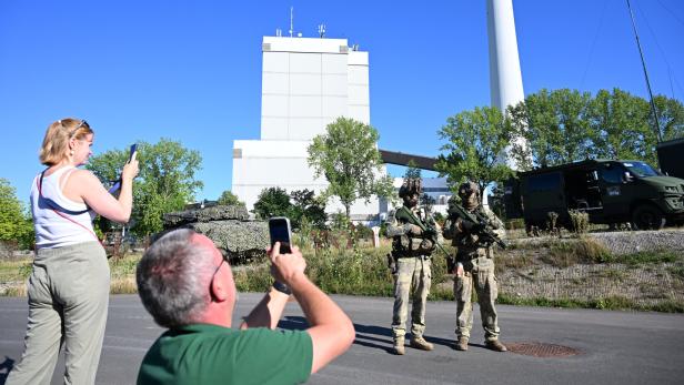 Zwei Personen fotografieren mit ihren Smartphones zwei schwer bewaffnete Soldaten in Tarnkleidung vor einem Kraftwerk.