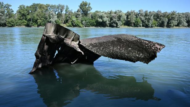Ein verrostetes Wrackteil ragt aus dem Wasser eines Flusses vor einem bewaldeten Ufer hervor.
