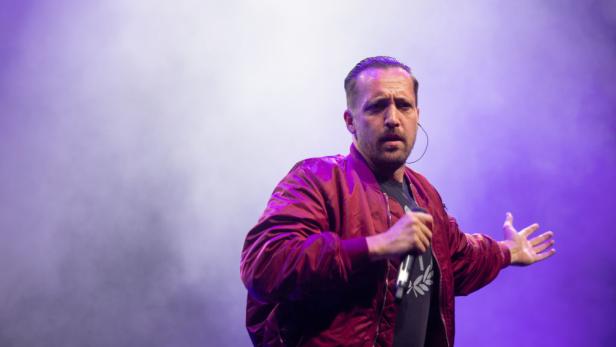 Danger Dan steht mit Mikrofon auf der Bühne. Er trägt eine rote Bomberjacke.