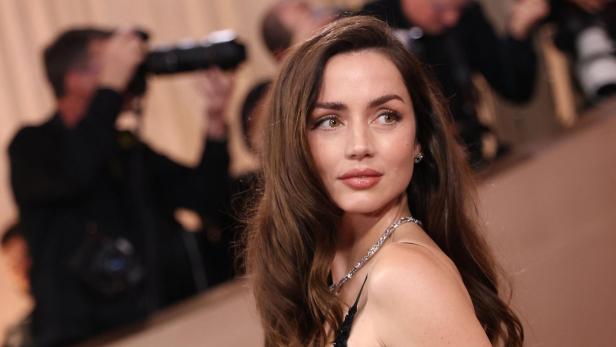 Ana de Armas blickt mit langem, braunem Haar und in einem schwarzen Abendkleid vor einer Kulisse mit Fotografen zur Seite.