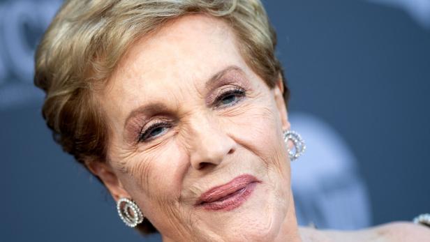 Nahaufnahme der Schauspielerin Julie Andrews mit kurzem blondem Haar und auffälligen Ohrringen vor einem dunklen Hintergrund.