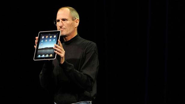 Steve Jobs präsentiert auf einer Bühne ein iPad vor schwarzem Hintergrund.