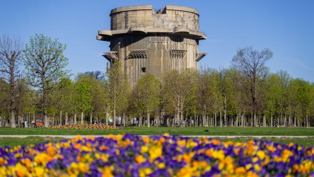 Der Wiener Flakturm im Augarten ragt hinter einer blühenden Blumenwiese und Bäumen in den blauen Himmel.