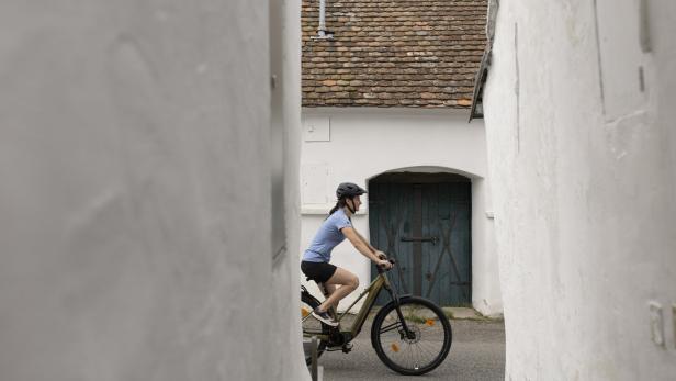 Fahrradfahrerin mit Helm fährt durch eine enge Gasse vor einem weißen Haus mit blauem Tor.