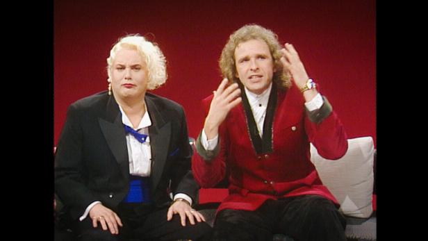 Hella von Sinnen und Thomas Gottschalk sitzen vor einem roten Hintergrund und unterhalten sich.