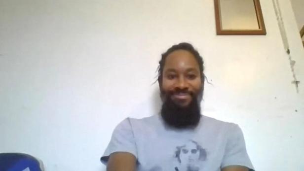 Ein lächelnder Mann mit Bart und Dreadlocks trägt ein graues T-Shirt vor einer weißen Wand.