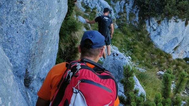 Zwei Wanderer klettern auf einem schmalen Bergpfad zwischen Felsen und Latschen.