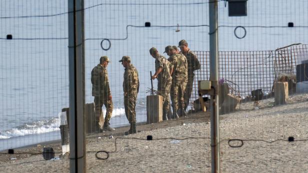 Sechs Soldaten in Tarnuniform stehen am Strand hinter einem Zaun, das Meer im Hintergrund.