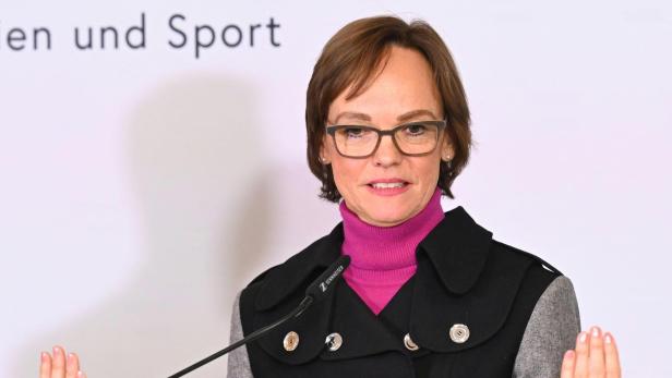 Frau mit Brille spricht am Mikrofon und hebt beide Hände vor einem hellen Hintergrund.