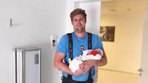 Feuerwehrmann in Einsatzkleidung hält sein neugeborenes Kind im Krankenhaus in den Armen.