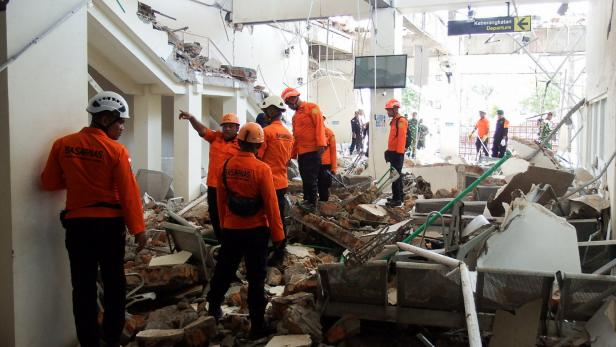 Rettungskräfte in Orange und Helmen inspizieren die Trümmerhalle eines teilweise eingestürzten Gebäudes.