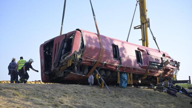 Ein Kran hebt einen verunglückten roten Reisebus von einer Böschung, während Einsatzkräfte daneben stehen.