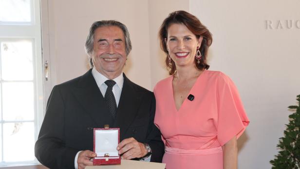 Riccardo Muti hält ein Schmuckkästchen mit einem Ring in der Hand und lächelt neben einer Frau in einem lachsfarbenen Kleid.