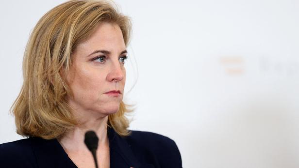 Eine blonde Frau im dunklen Blazer spricht an einem Mikrofon vor hellem Hintergrund.